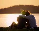 Ein Paar sitzt am Seeufer im Sonnenuntergang, die Arme umeinander geschlungen. Sie finden Trost und lernen, sich selbst zu verzeihen.