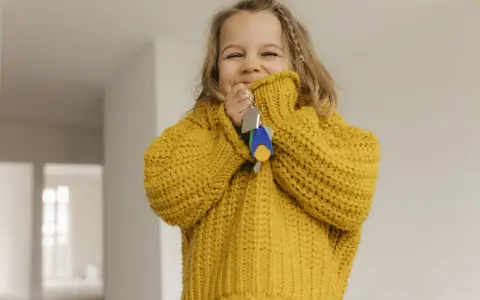 Ein fröhliches kleines Mädchen im gelben Pullover hält Schlüssel in der Hand. Die Frage "ab wann Kinder alleine lassen" beschäftigt Eltern oft, doch dieses Bild strahlt Unbeschwertheit aus.