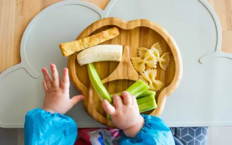 Babyhände greifen nach leckeren Baby-Snacks ab 6 Monaten: Banane, Gurke, Nudeln und ein Stück Brot auf einem Holztablett.