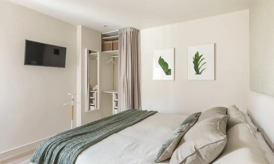 Ein offener Kleiderschrank, der wie ein Kleiderschrank selber bauen mit Vorhang wirkt, mit Regalen und Kleiderstange. Daneben ein Spiegel und ein Bett mit grüner Decke.