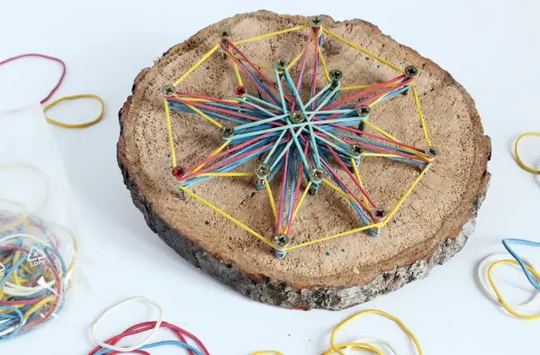 Bunte Gummibänder spannen sich zu einem Stern auf einer Baumscheibe. Ein tolles Bastelprojekt für Kinder, das Kreativität und Feinmotorik fördert.