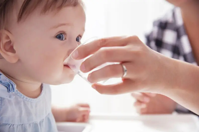 Ein Baby trinkt vorsichtig Wasser aus einem Becher, gehalten von einer Hand. Die Frage "ab wann Baby Wasser geben?" wird hier visuell beantwortet.